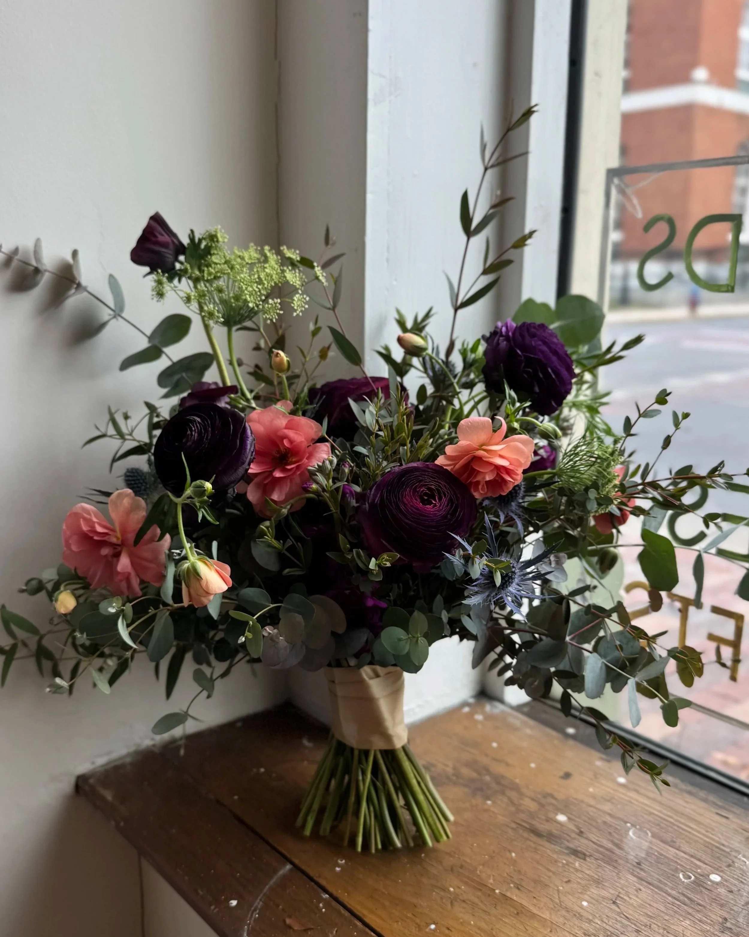 A flower bouquet with purple, pink, and green flowers and foliage, wrapped with beige paper, placed on a wooden surface next to a window.