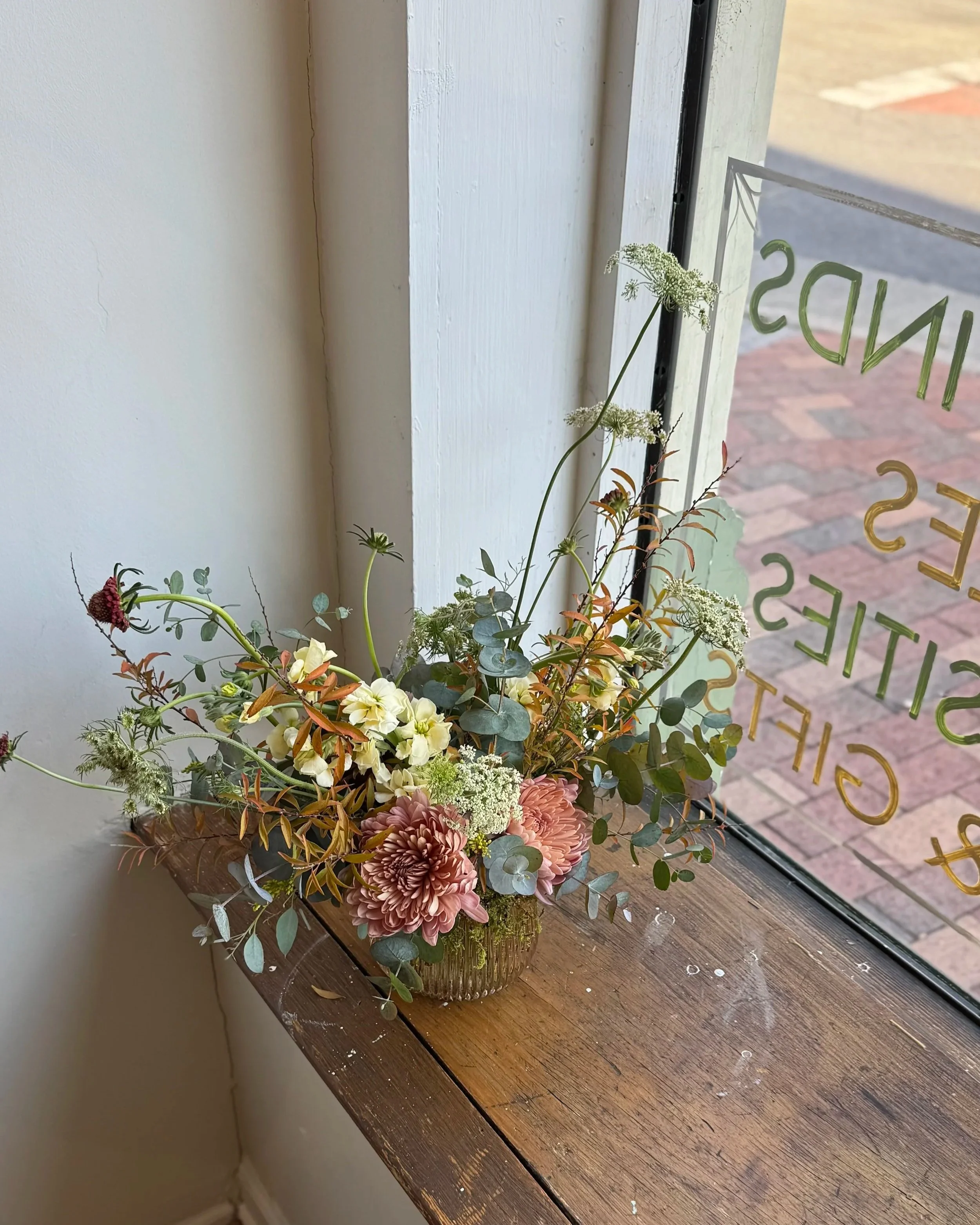 A floral arrangement in a glass vase on a wooden windowsill, near a glass door with lettering, featuring pink dahlias, white flowers, green eucalyptus leaves, and other greenery.