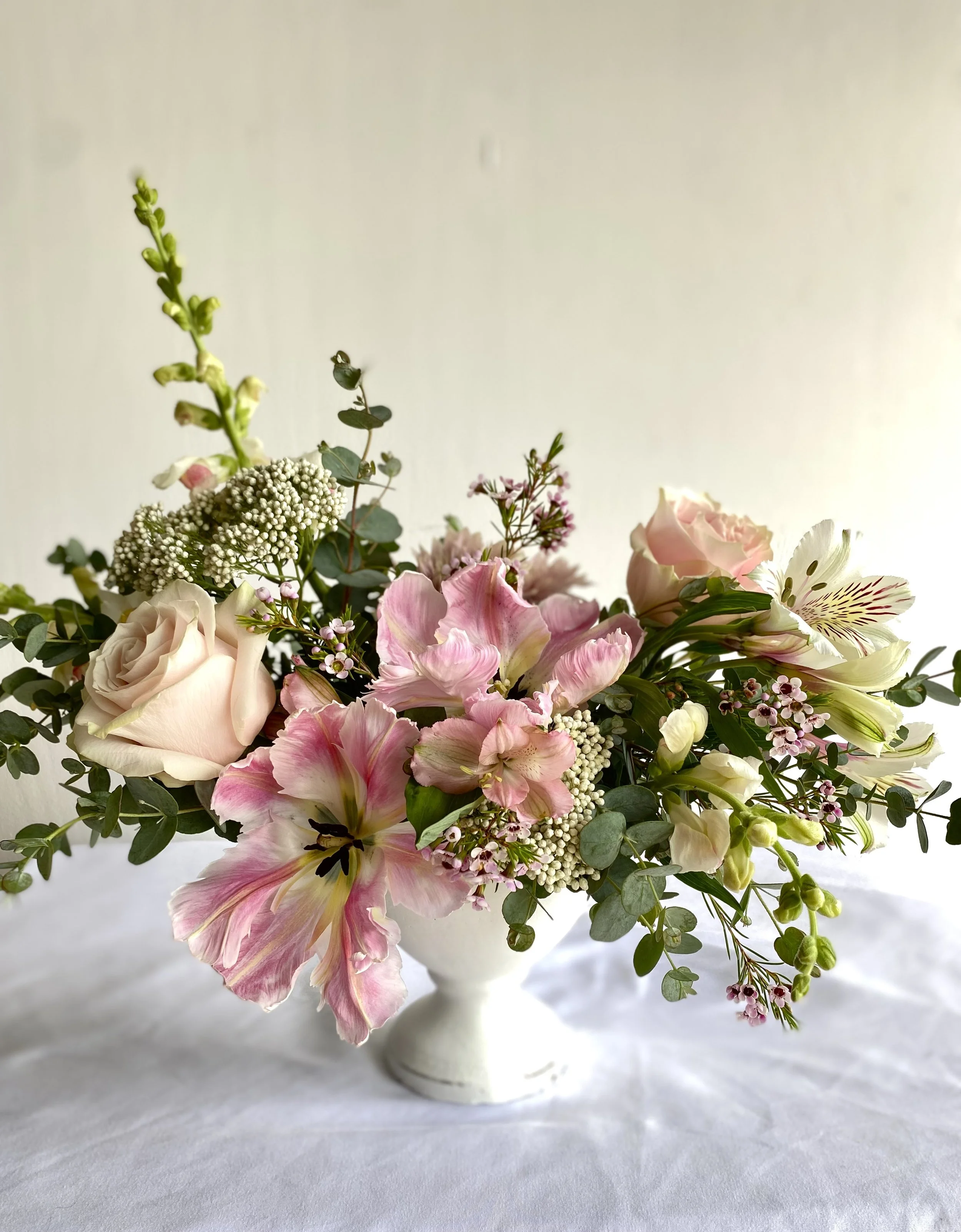 A floral arrangement in a white vase with pink and white flowers and green foliage on a white surface.
