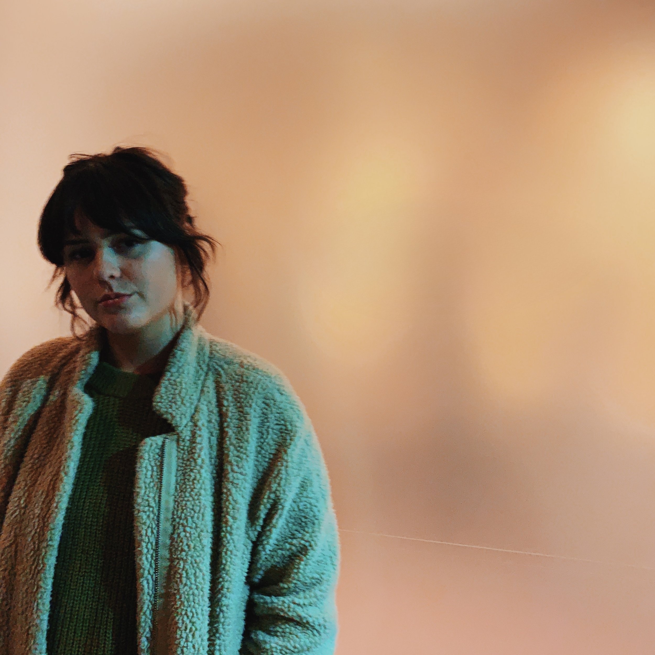 A woman with dark, wavy hair wearing a tan fleece jacket and green top, standing against a plain peach-colored wall.