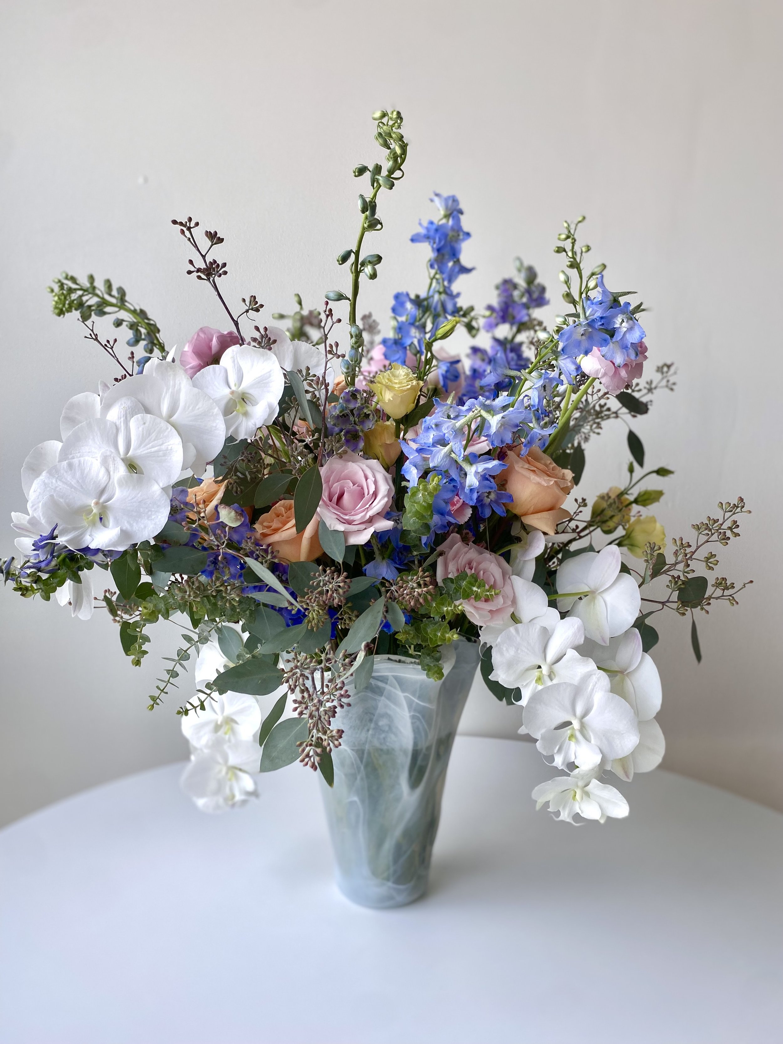 A bouquet of mixed flowers in a marble pattern vase on a white table, featuring white orchids, pink roses, blue delphiniums, and greenery.