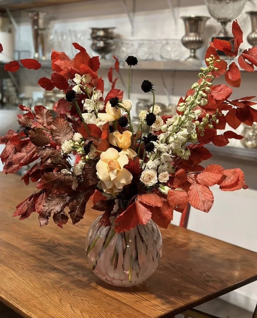A floral arrangement with red leaves, white, cream, and yellow flowers in a transparent glass vase on a wooden table.