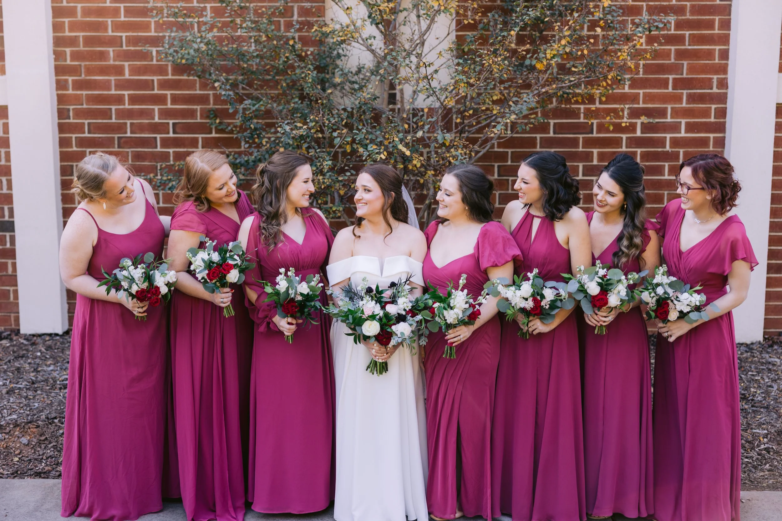 Bridal party of nine women standing outdoors in front of a brick wall, wearing matching magenta dresses, holding bouquets, with the bride in a white dress in the center.