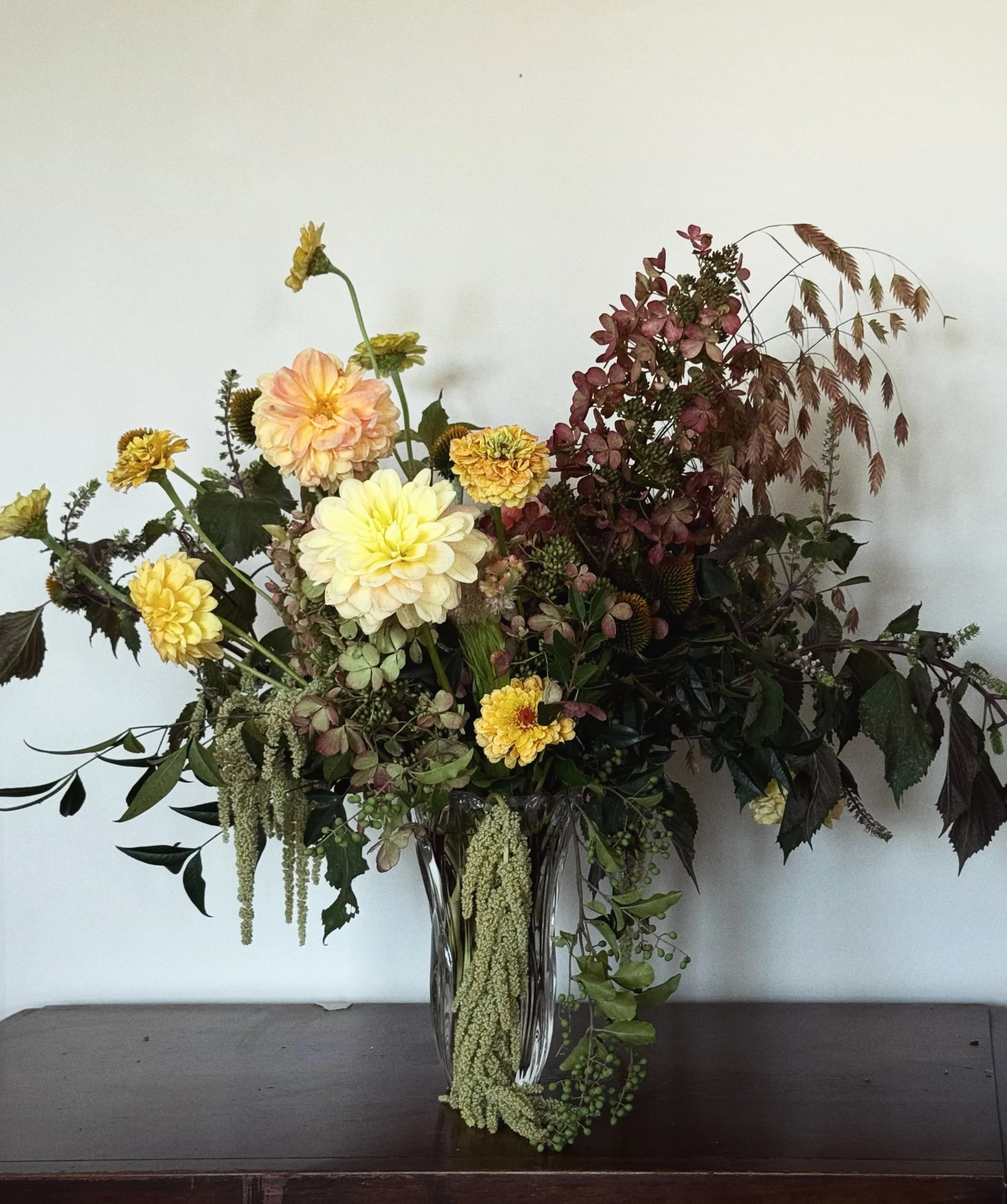 Arrangement of yellow and peach flowers with green and brown foliage in a clear glass vase on a wooden surface against a white wall.