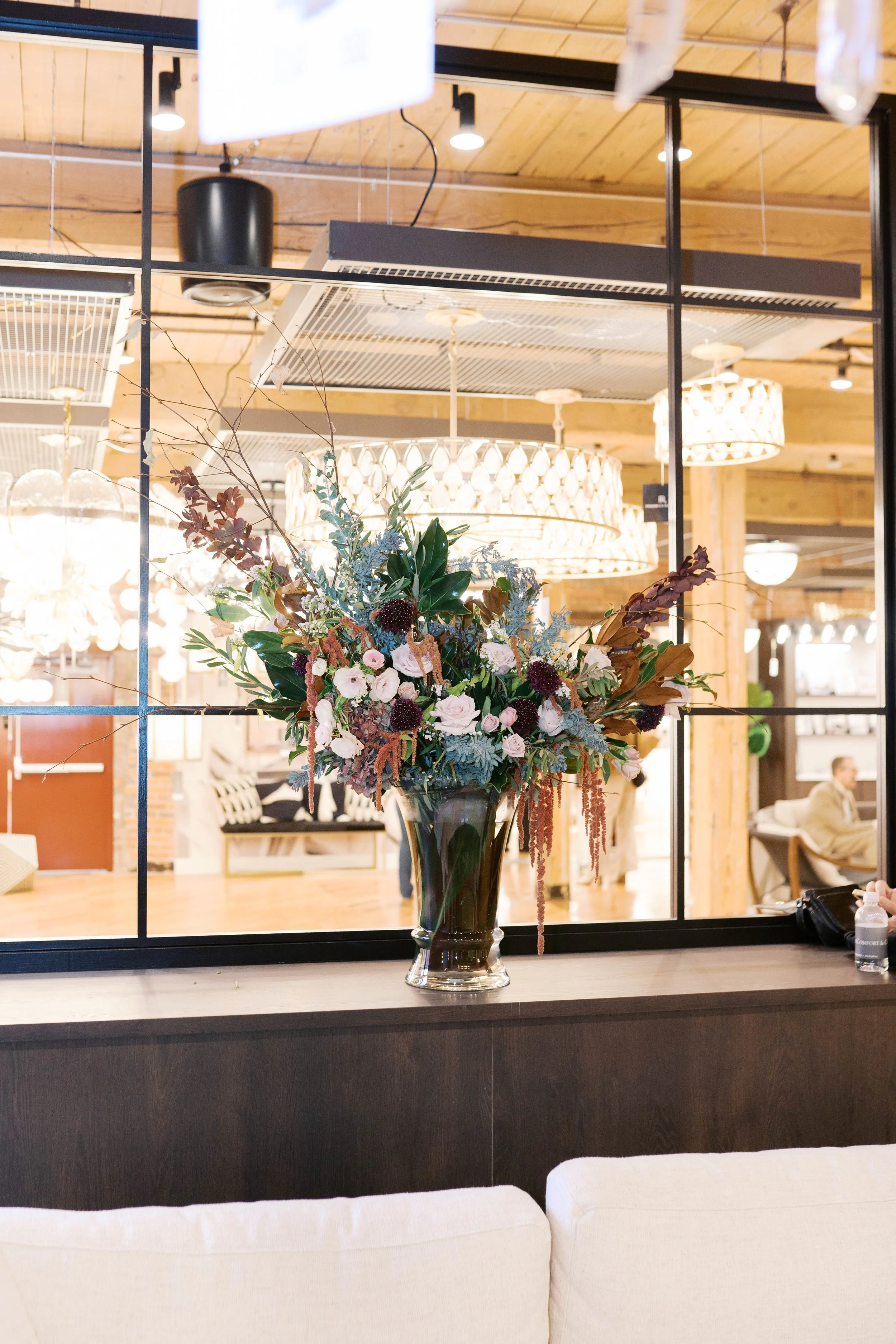 A large floral arrangement in a glass vase on a dark wooden ledge, seen through a black metal window frame. The background features a warmly lit interior with wooden ceiling, hanging chandeliers, and people sitting at tables.