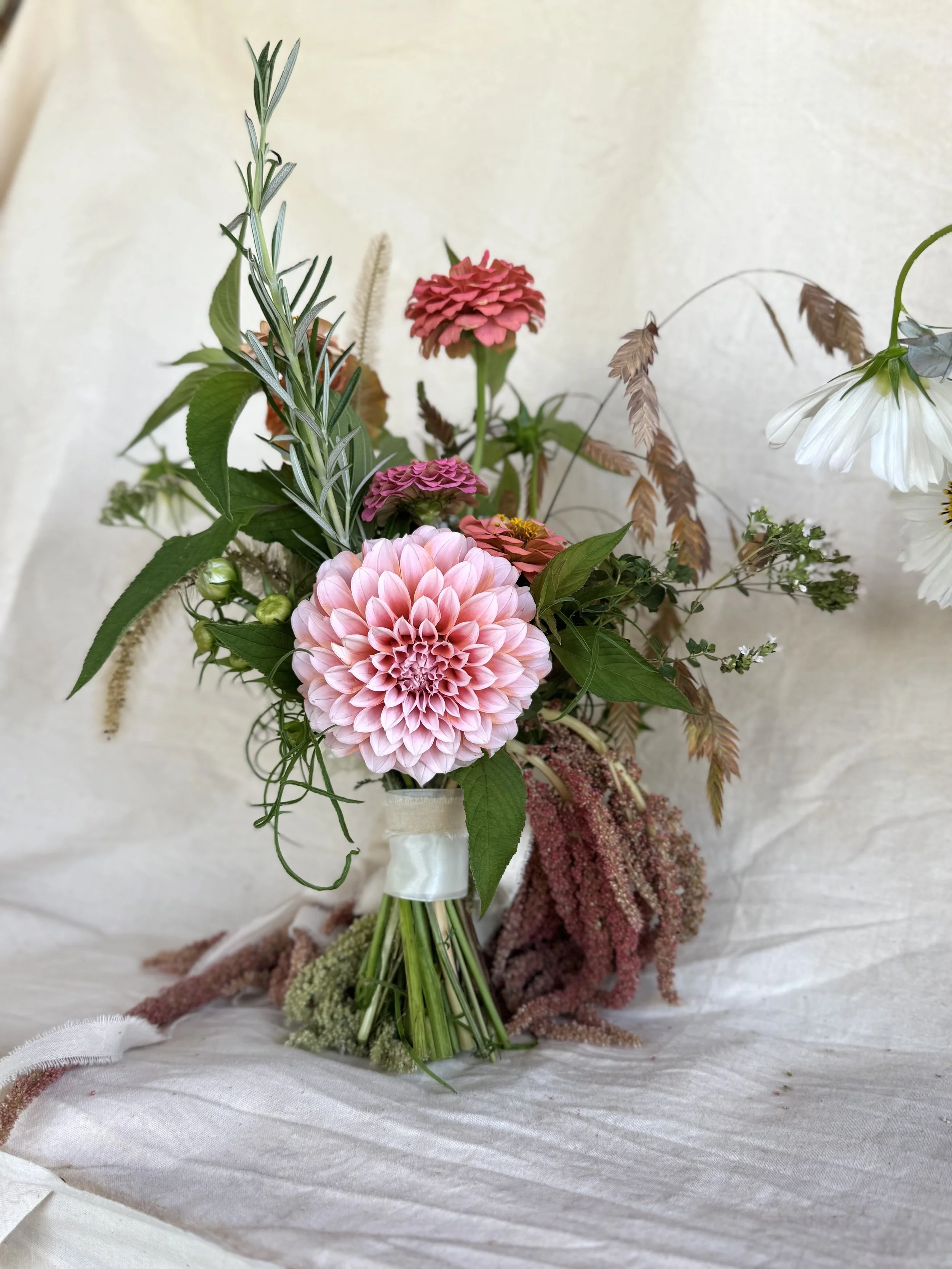 A bouquet of various flowers including a large pink dahlia, pink zinnias, white cosmos, and greenery, arranged in a small glass vase on a white fabric surface.