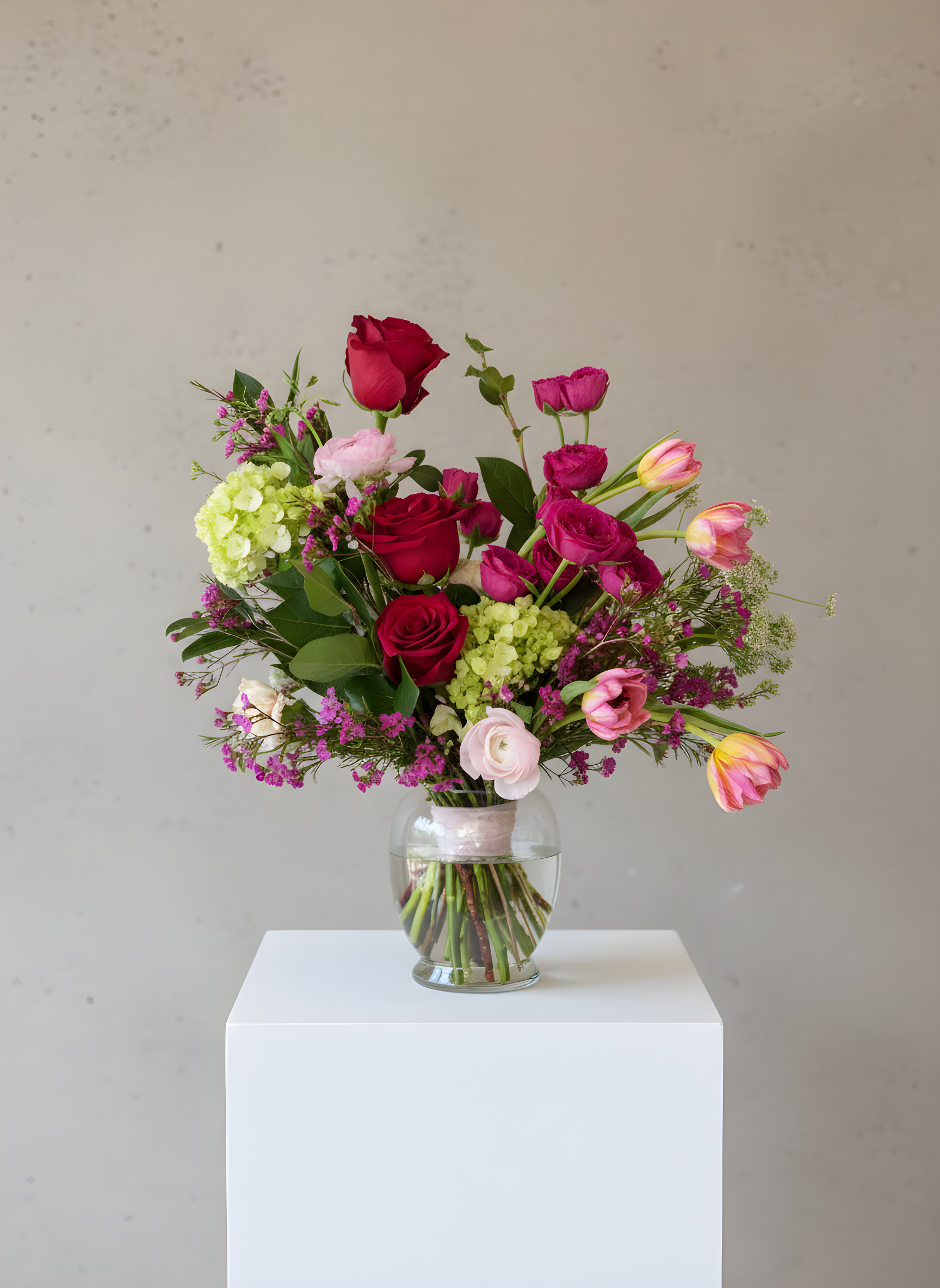 A colorful bouquet of flowers in a glass vase on a white pedestal, featuring roses, tulips, and assorted blooms.