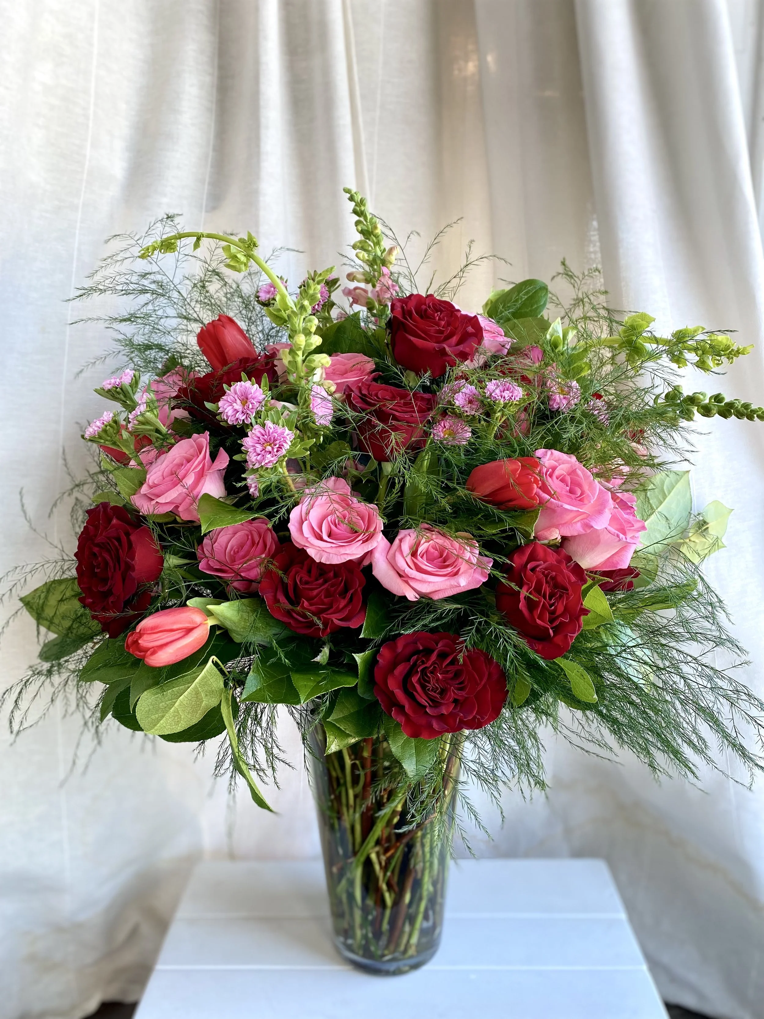 A tall glass vase filled with pink and red roses, tulips, and assorted greenery.