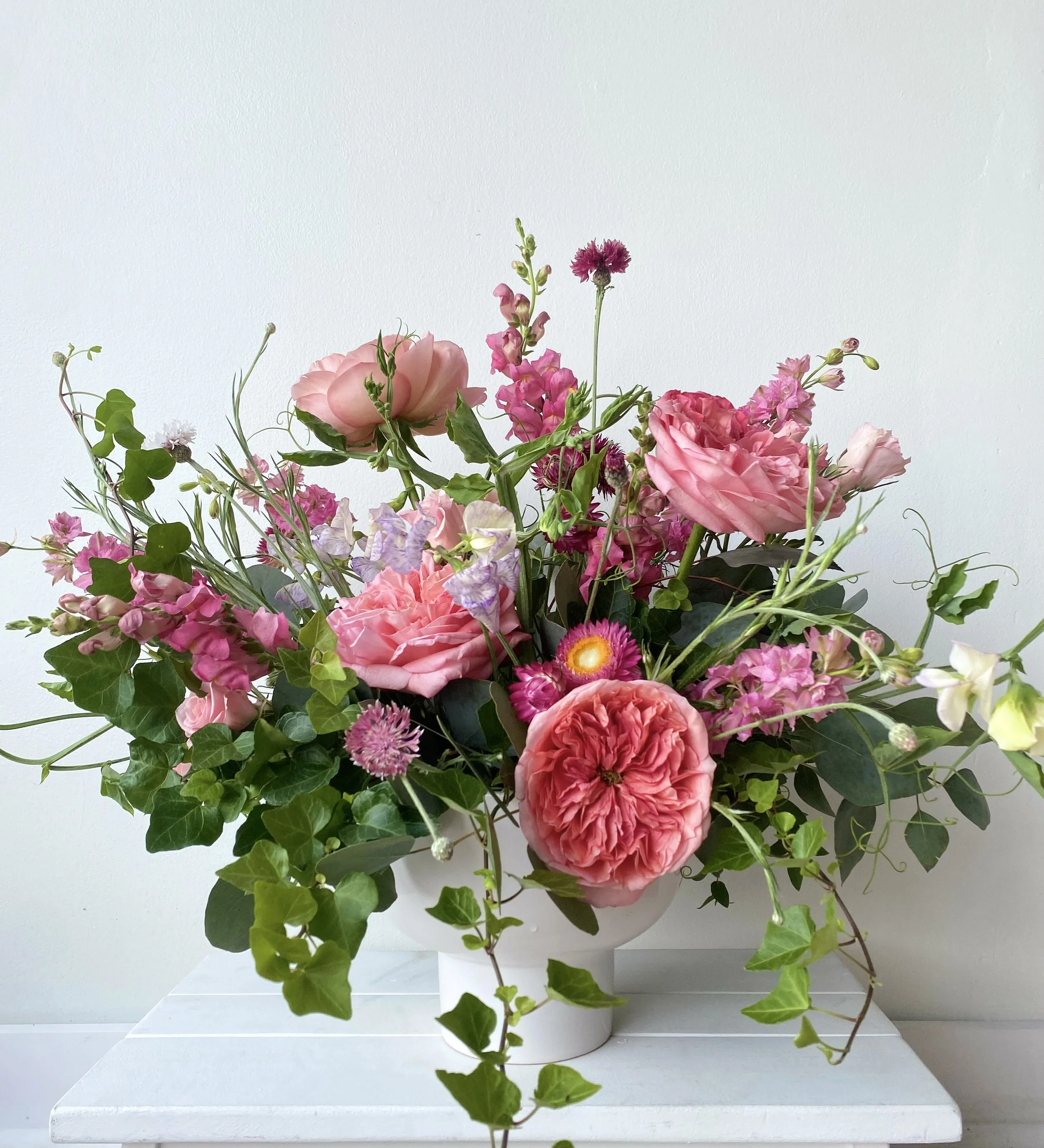 Pink and purple mixed flower bouquet in a white vase on a white surface against a plain white background.