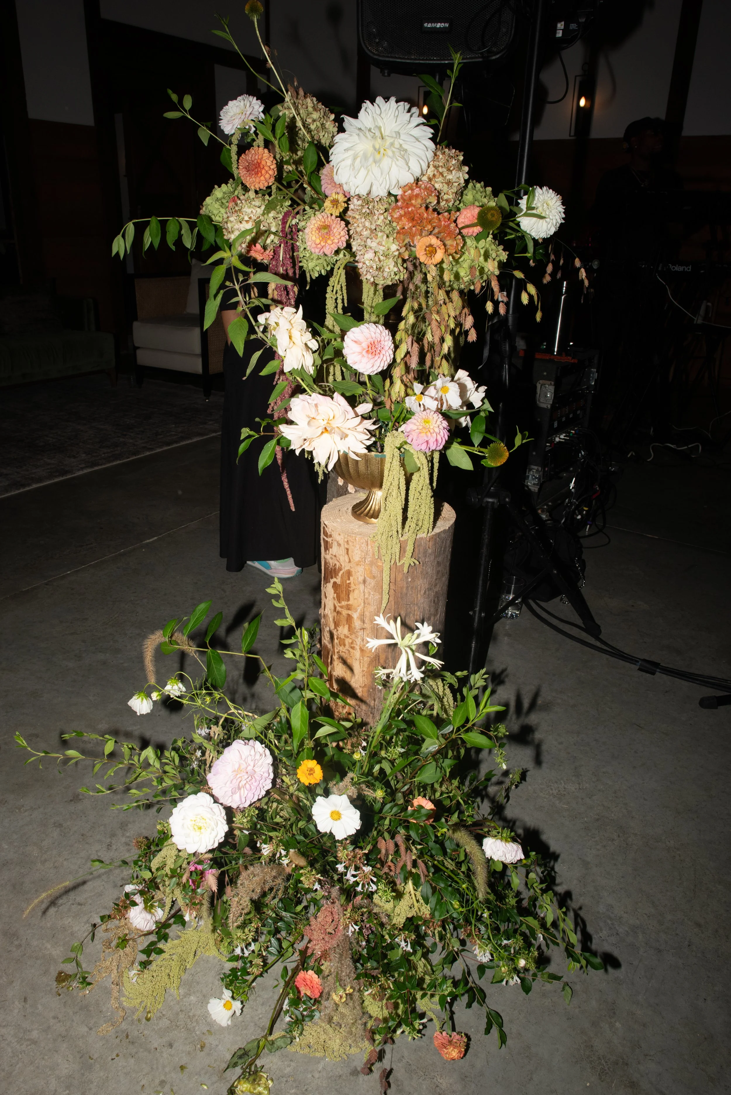 A large floral arrangement with a variety of flowers including peonies and dahlias, in a gold vase atop a wooden log, with a second, smaller floral bouquet on the floor in front of it.