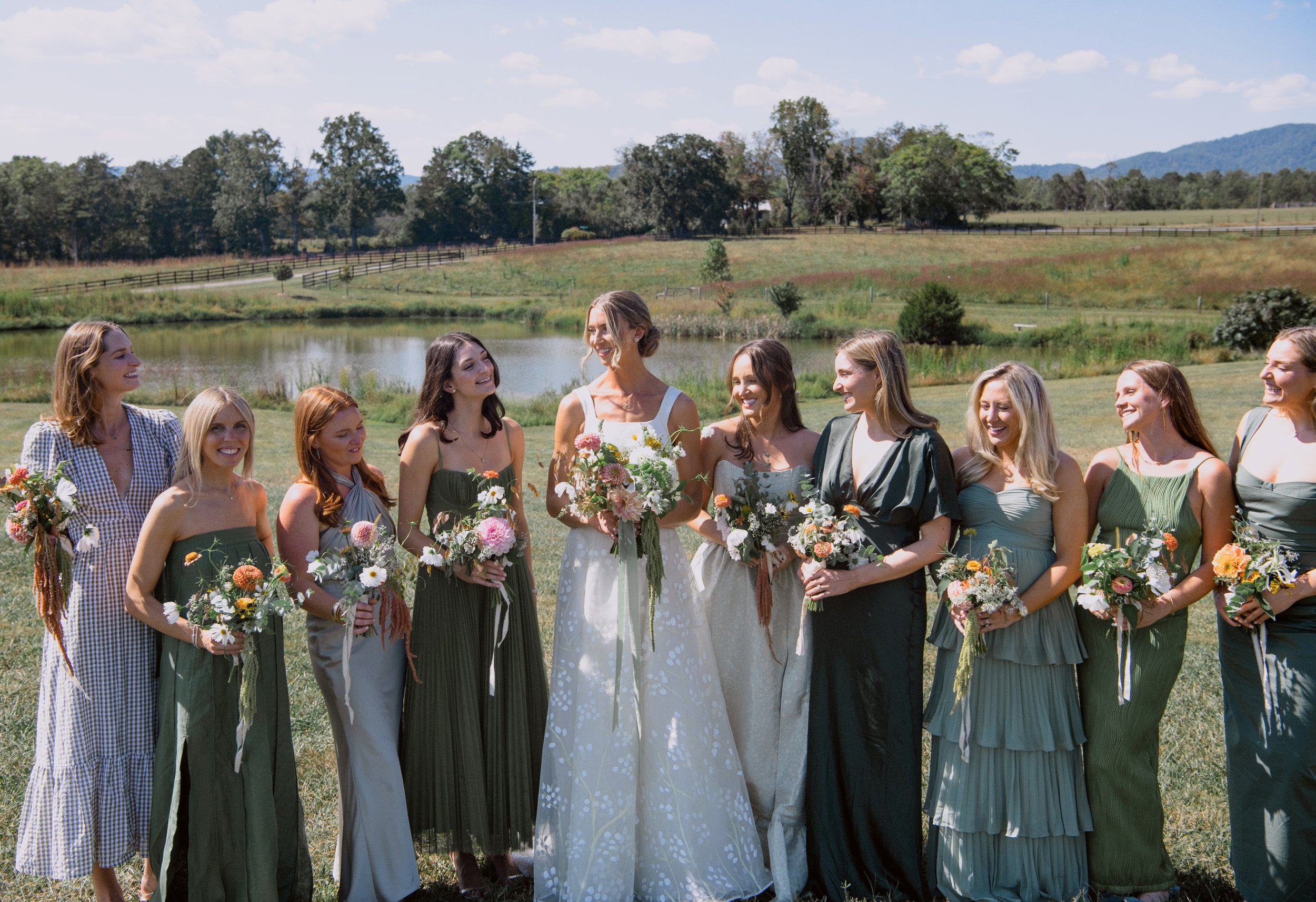 A group of women in colorful dresses standing outdoors by a pond with trees and hills in the background, holding bouquets of flowers, during a sunny day.