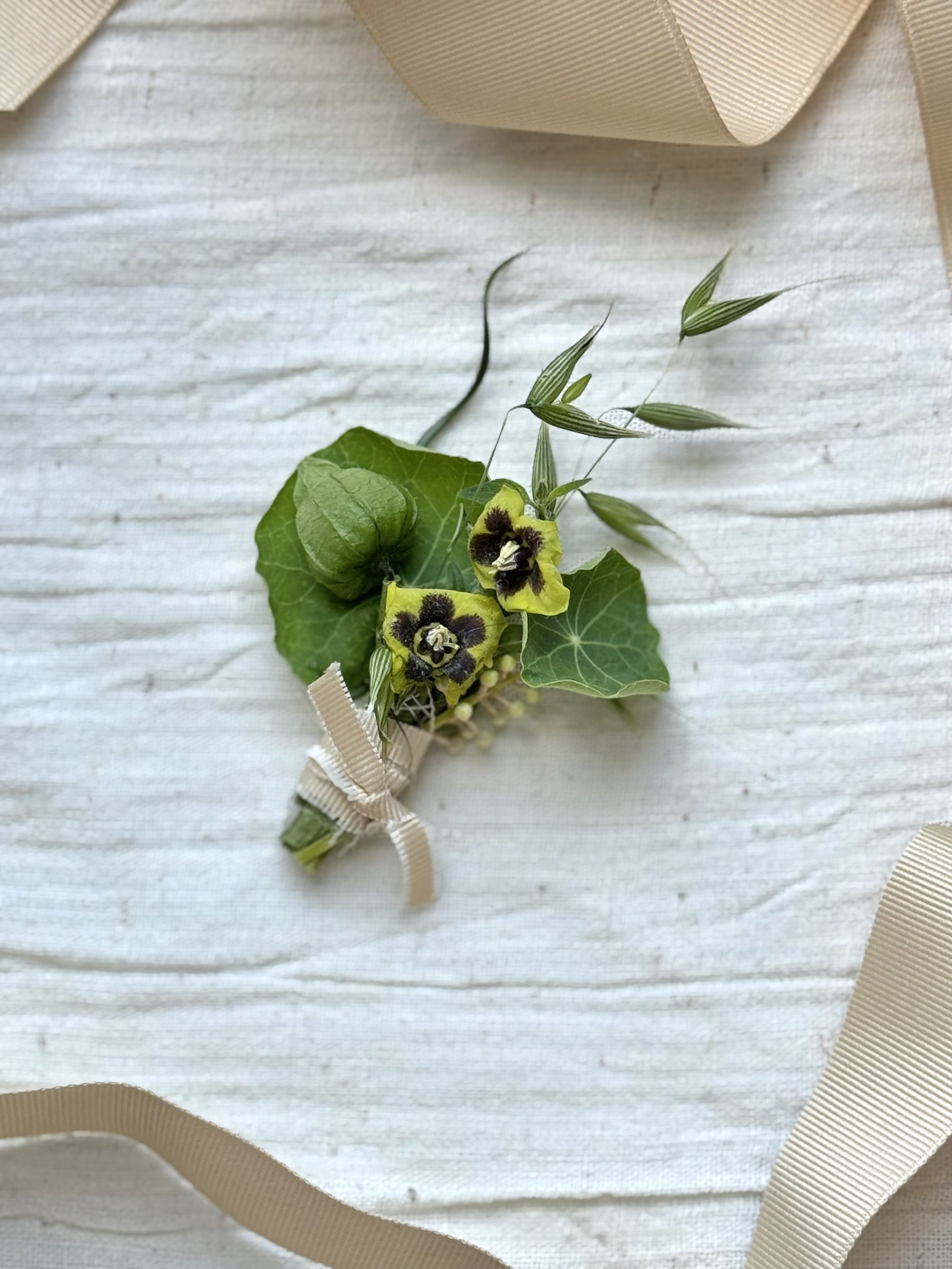A small boutonniere with green leaves, yellow and black flowers, and beige ribbon, resting on a white wooden surface with a beige ribbon nearby.