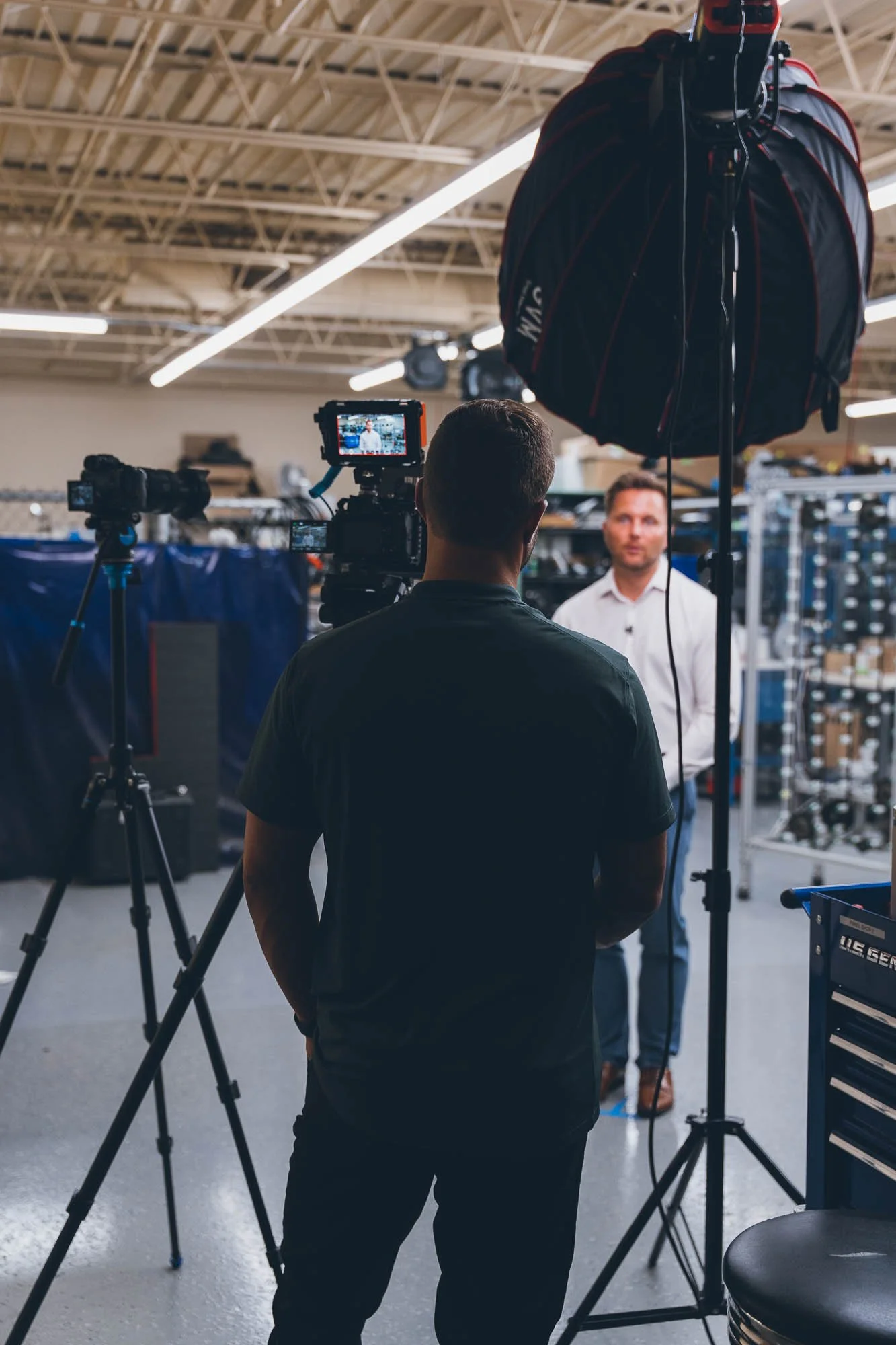A man is filming a person being interviewed in an industrial workspace, with professional lighting and camera equipment around.