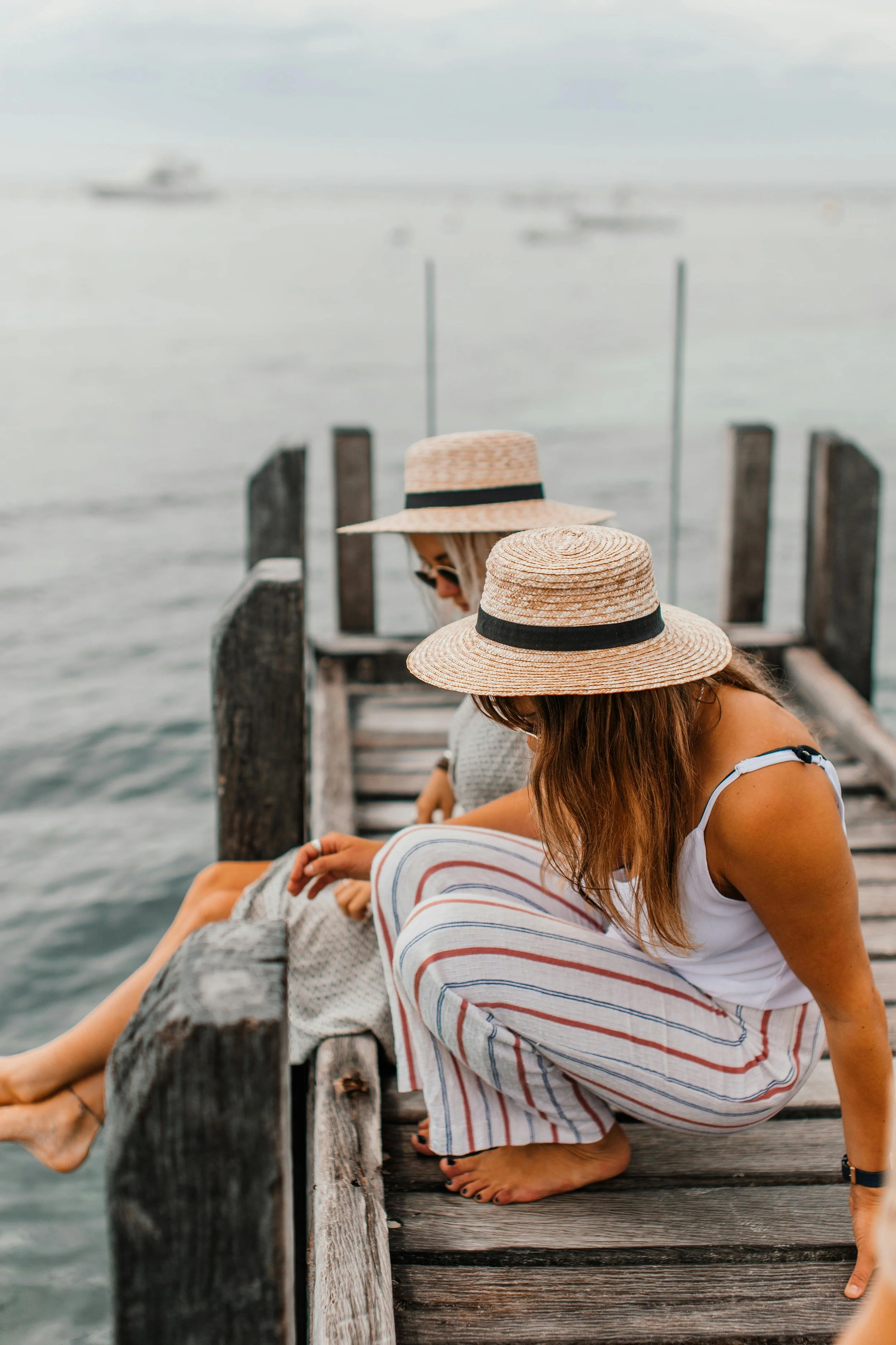 Two women wearing wide-brimmed hats sitting on a wooden dock by the water, with boats visible in the background.