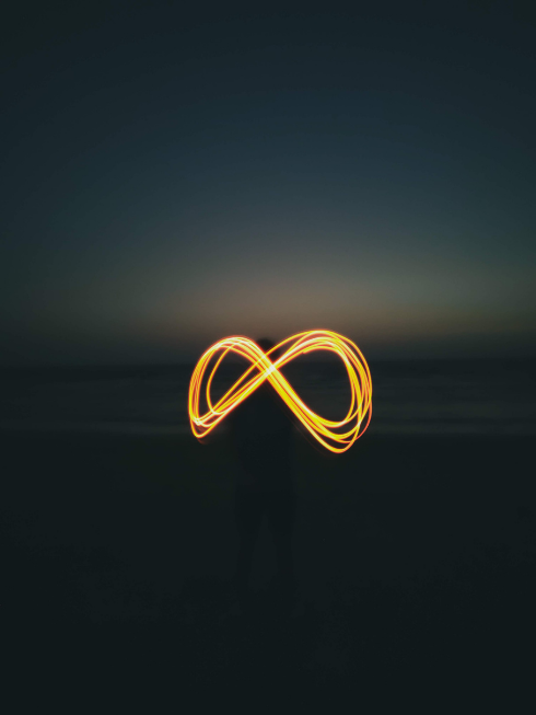 Person standing on a beach at twilight holding a light source creating an infinity symbol with yellow and orange light trails.