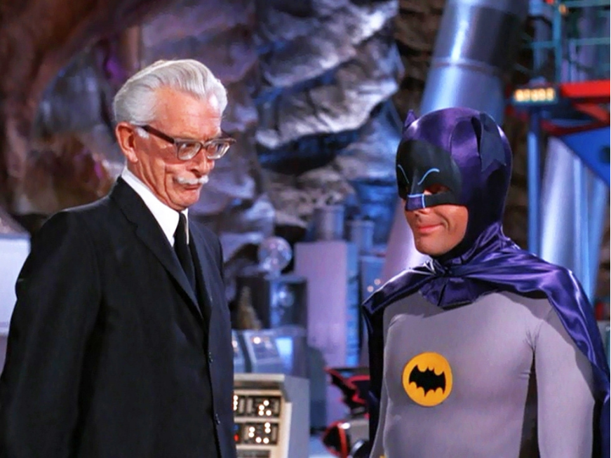 Two men dressed as Batman and Alfred Pennyworth from the Batman TV series, standing and smiling inside the Batcave.