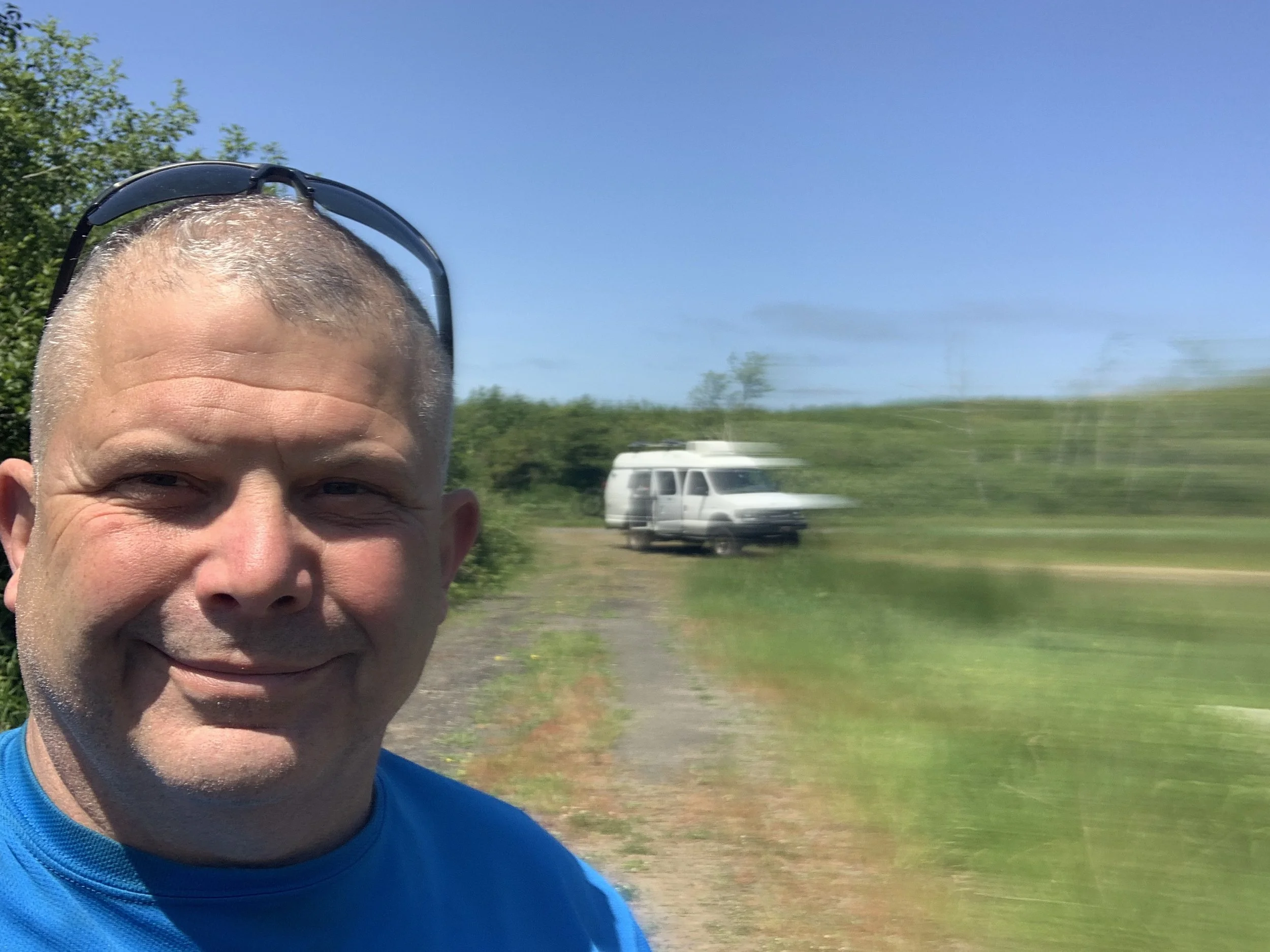 A man with short gray hair, wearing glasses on his head and a blue shirt, smiling in a rural outdoor setting with a blurred white van in the background on a sunny day.