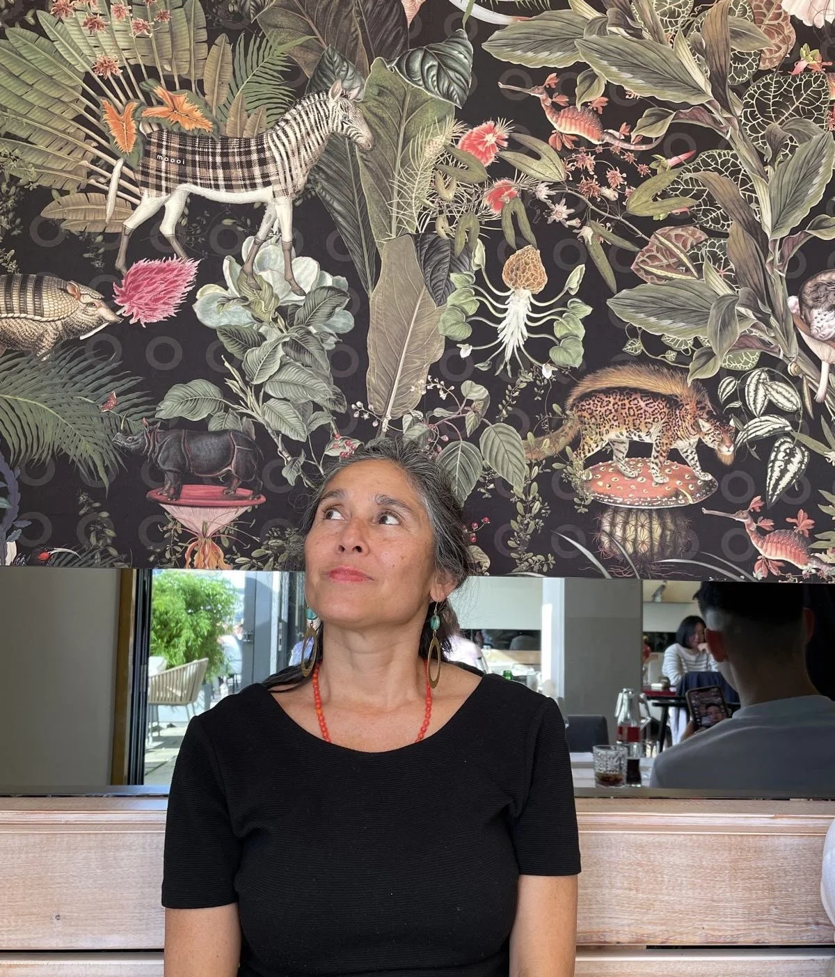 Ein Porträt einer Frau mit dunklem Haar, die in einem Café sitzt. Über ihr hängt eine auffällige Wandtapete mit einem Dschungel-Design, das verschiedene Tiere wie Leoparden, Vögel und andere Kreaturen zeigt, umgeben von großen Blättern und Pflanzen.