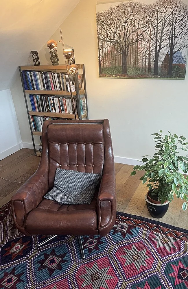 A cozy corner with a brown leather armchair with a checkered pillow, a tall bookshelf filled with books, a potted plant, a colorful patterned rug, a modern floor lamp, and a large painting of a leafless tree on the wall.