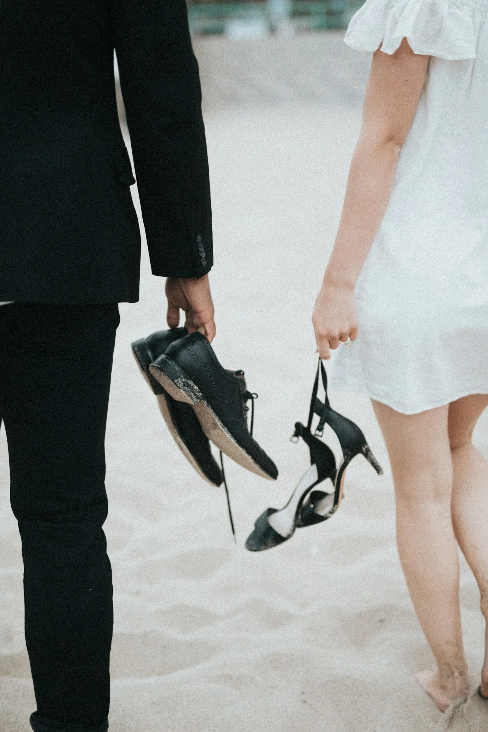 A man in a black suit holding black dress shoes, and a woman in a white dress holding black and white high-heeled shoes, standing on sandy ground.