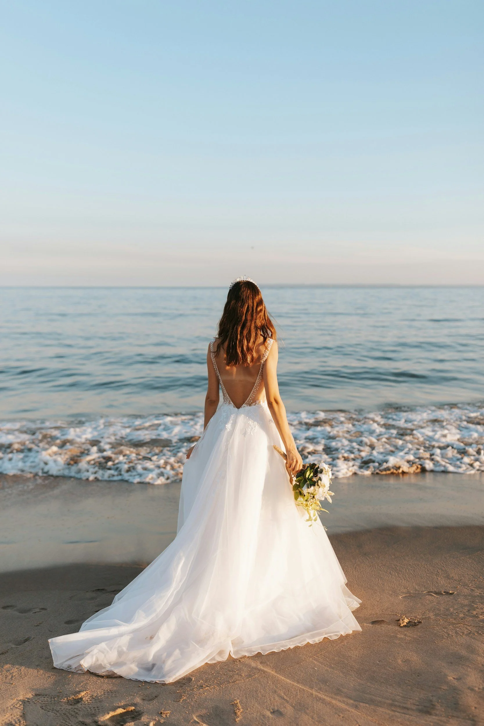 A woman in a wedding dress holding a bouquet stands on the beach facing the ocean during sunset.