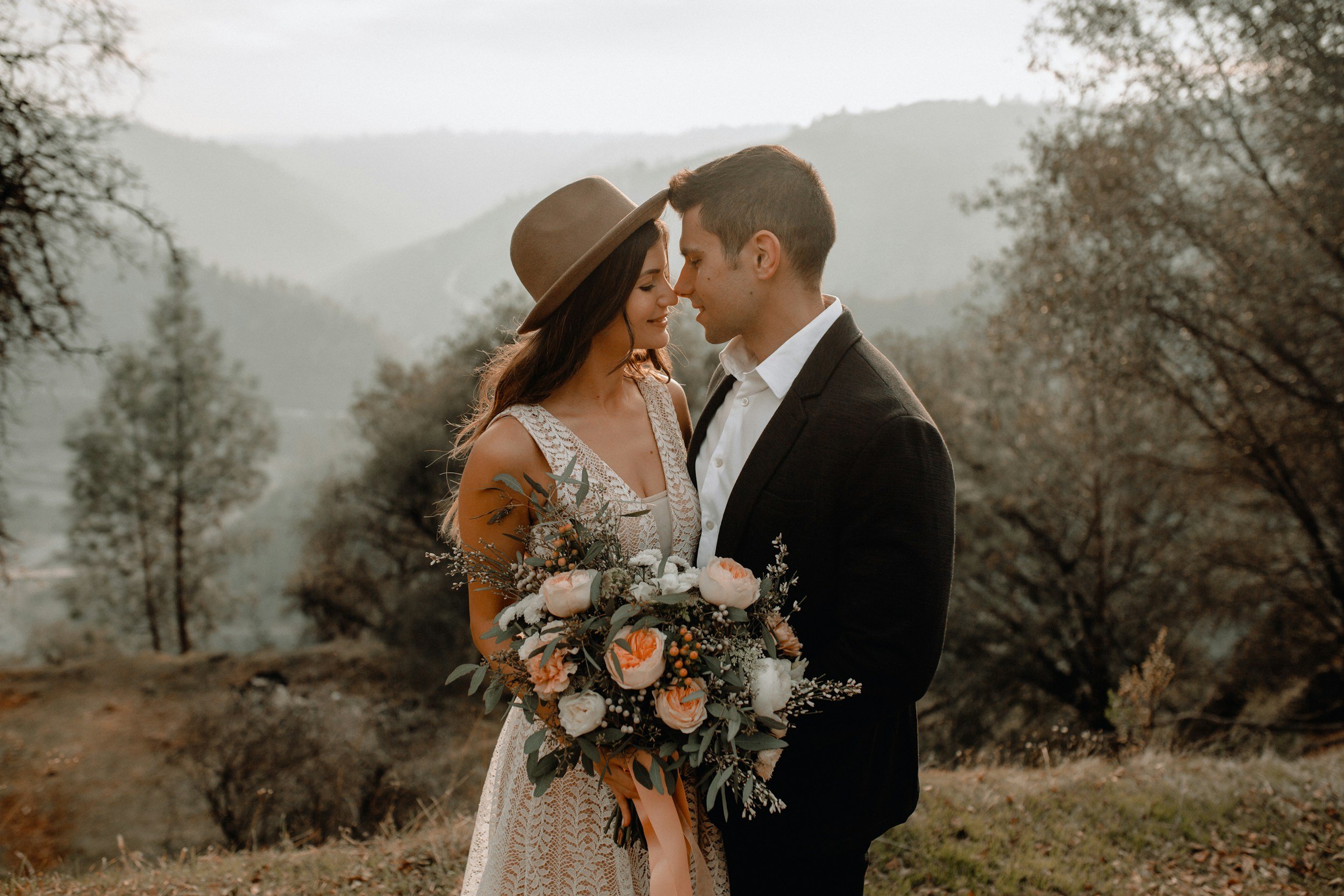 A woman and man stand close together in an outdoor setting with trees and mountains, gazing at each other. The woman is wearing a lace dress, a hat, and holding a bouquet of flowers. The man is wearing a dark suit and white shirt.