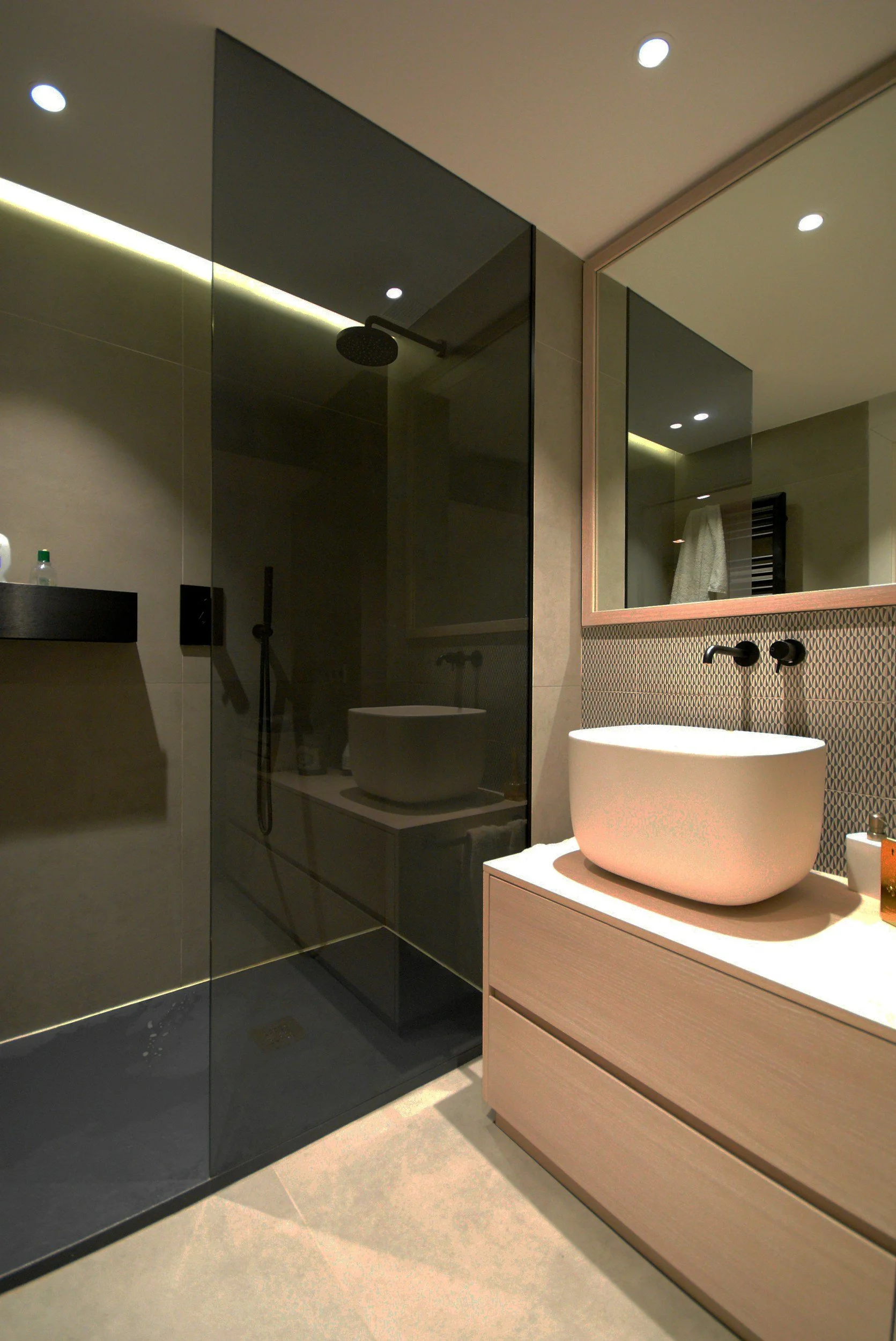 Modern bathroom with a walk-in shower with black glass doors, a large rectangular mirror, a vessel sink on a light wooden vanity, and tiled walls and floor.