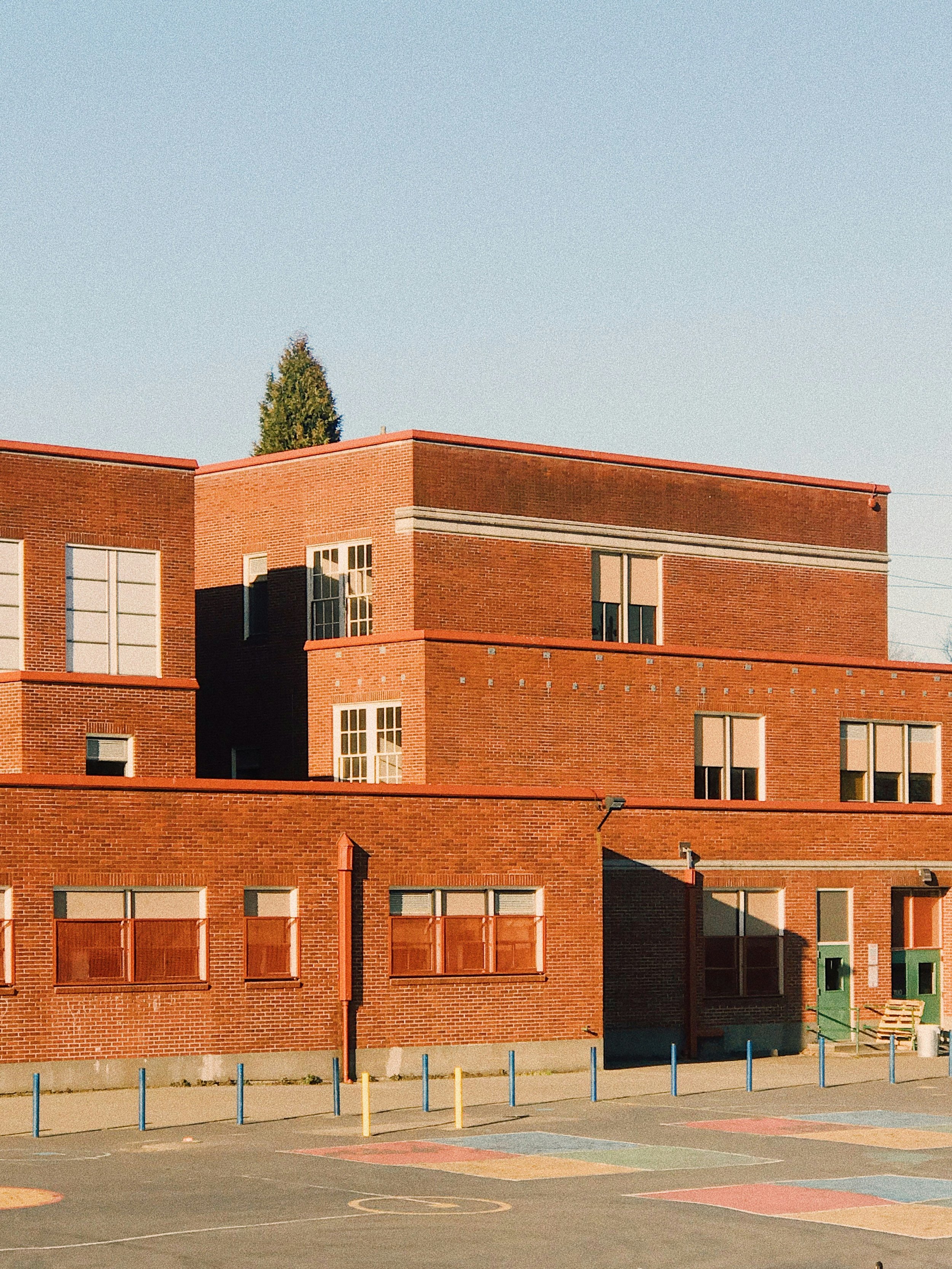 A brick school building with multiple floors and windows, adjacent to a parking lot with colorful painted basketball court lines and blue and yellow posts, green trees in the background, under a clear blue sky.