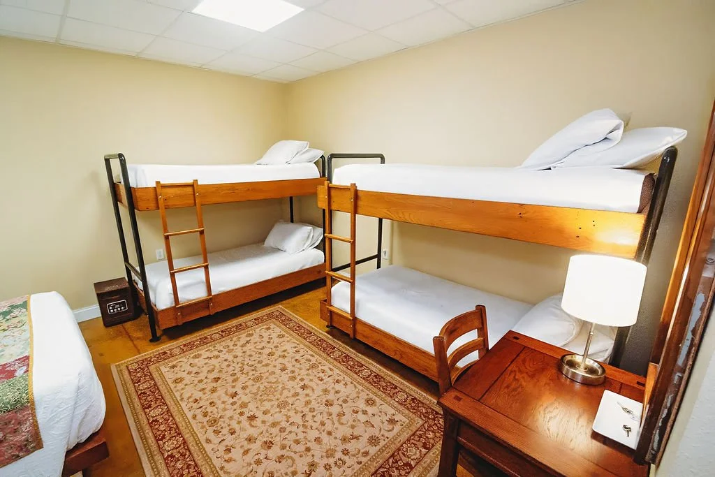 Room with two sets of wooden bunk beds, each with a pillow and white bedding, a small wooden desk with a lamp, a chair, and a patterned area rug.