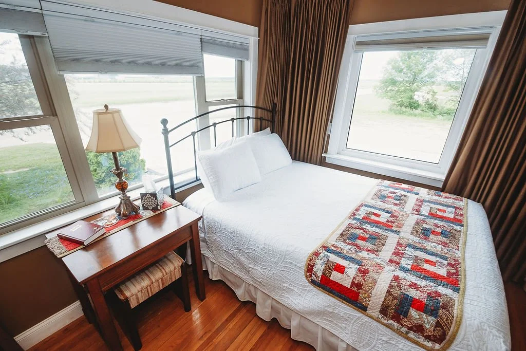 A cozy bedroom with a double bed covered in white bedding, a patchwork quilt at the foot, a wooden side table with a lamp, books, and decorative items, and large windows with brown curtains overlooking a green outdoor landscape.