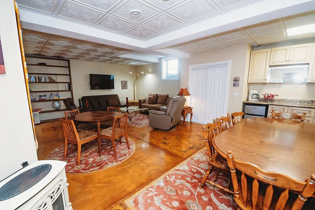 Living room with a dining table, living area with sofas, a wall-mounted TV, bookshelf, and a small kitchenette with a countertop and cabinets.