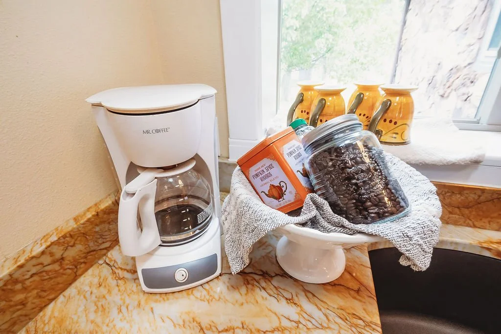 Coffee maker on a kitchen countertop next to a cake stand holding jars of baking supplies and a jar of coffee beans, with three ceramic mugs on the window sill in the background.