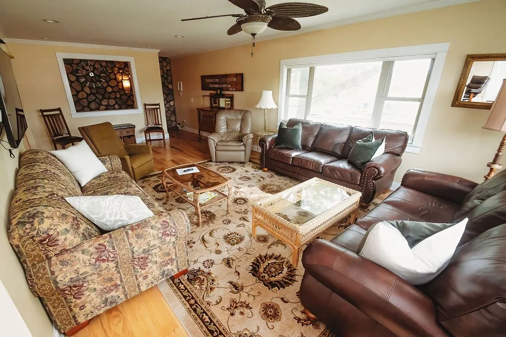 Living room with three sofas, two are leather and one has floral upholstery, a wooden coffee table, and a patterned area rug. Large window, ceiling fan, and a wall-mounted TV are also visible.