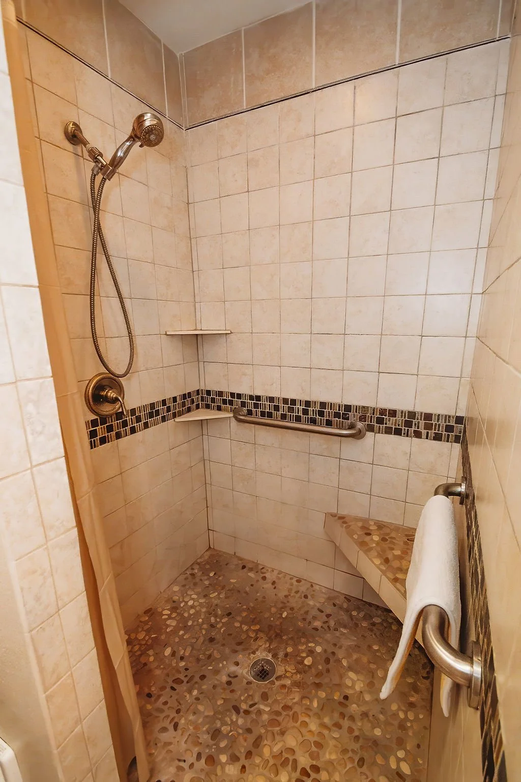 Walk-in shower with beige tiles, a mosaic tile stripe, a pebble floor, a built-in bench, a handheld showerhead, and a towel bar with a white towel.
