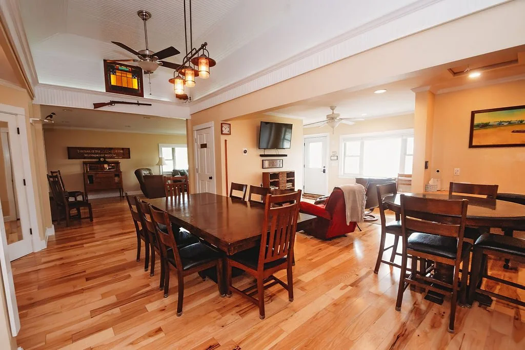 Open-concept living and dining room with wooden floors, a dining table with eight chairs, a sitting area with a red armchair and a brown sofa, a large flat-screen TV mounted on the wall, and bright windows letting in natural light.