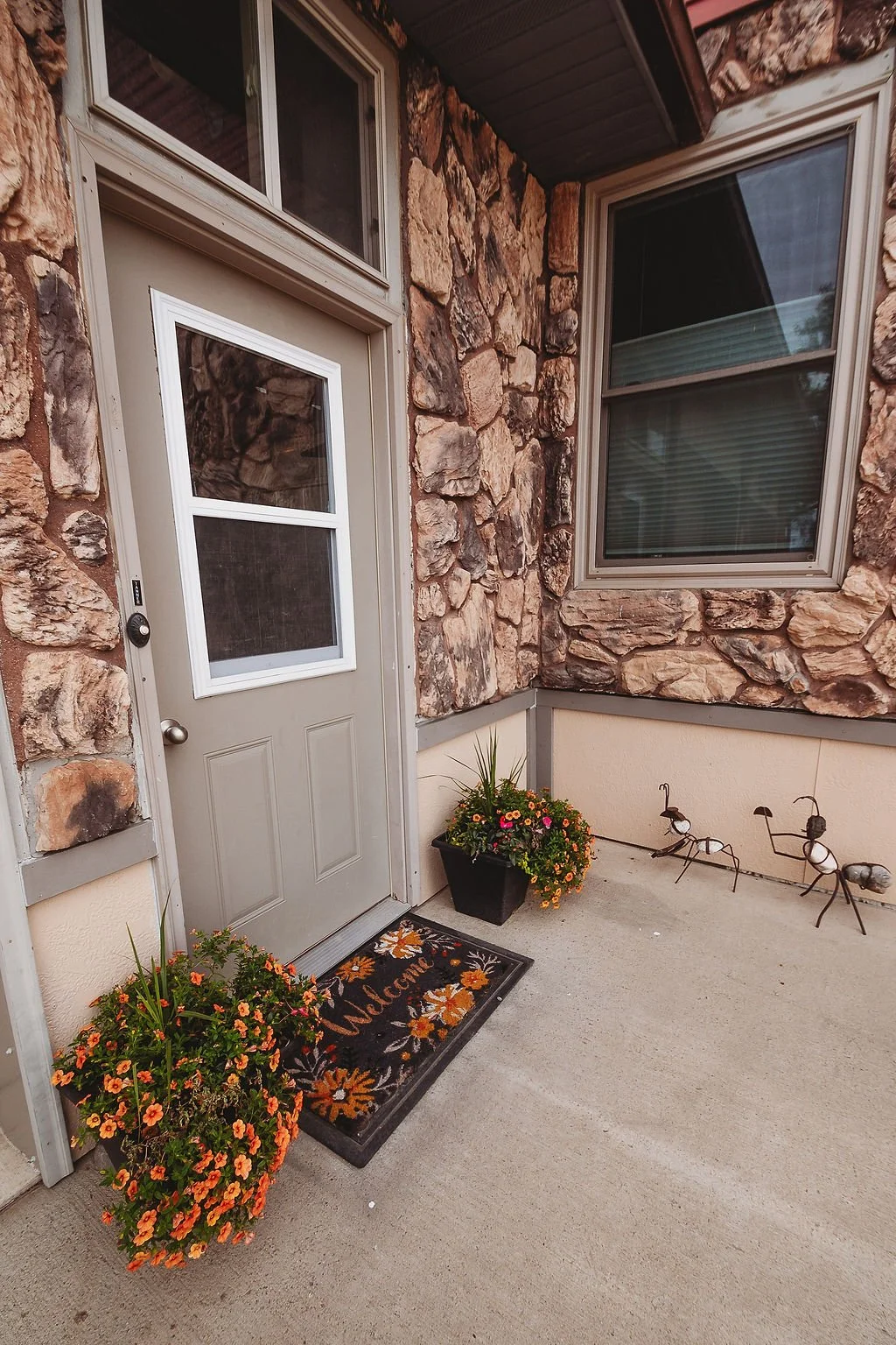 Front porch with a grey door, large stone and stucco exterior wall, two flower pots with colorful flowers, and decorative ant sculptures on the ground.