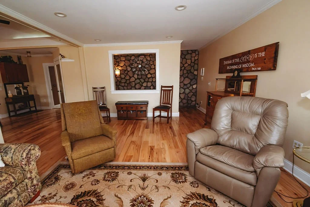Living room with beige walls, hardwood floors, a large beige leather armchair, a patterned fabric armchair, a wooden side table, and a decorative wall sign with a biblical quote. There are two wooden chairs and a small bench in the background, with a