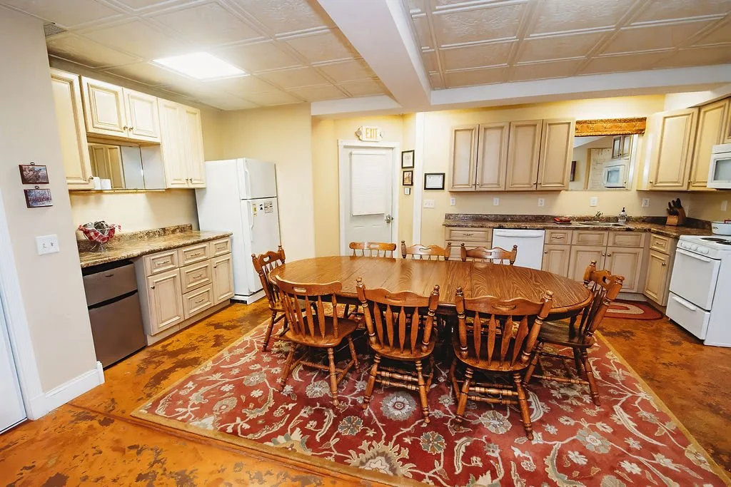 Kitchen with wooden dining table and eight chairs, beige kitchen cabinets, white appliances, and a large red patterned rug on hardwood flooring.