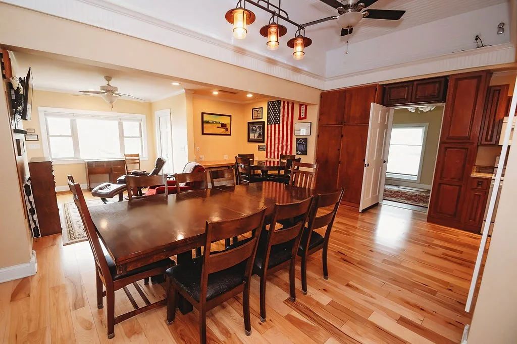 Open concept dining and living area with wooden dining table and chairs, exposed wooden cabinetry, American flag wall hanging, and large windows in the background.