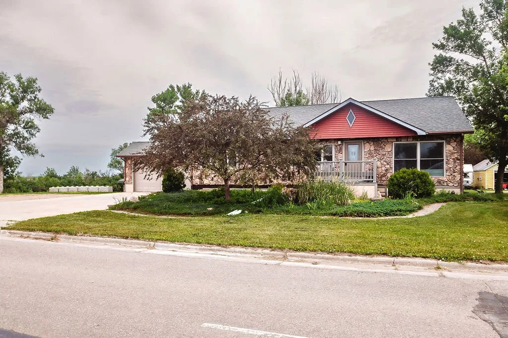 A single-story suburban house with a stone and red siding exterior, surrounded by a well-maintained lawn and trees, with a driveway on the left side, under a cloudy sky.