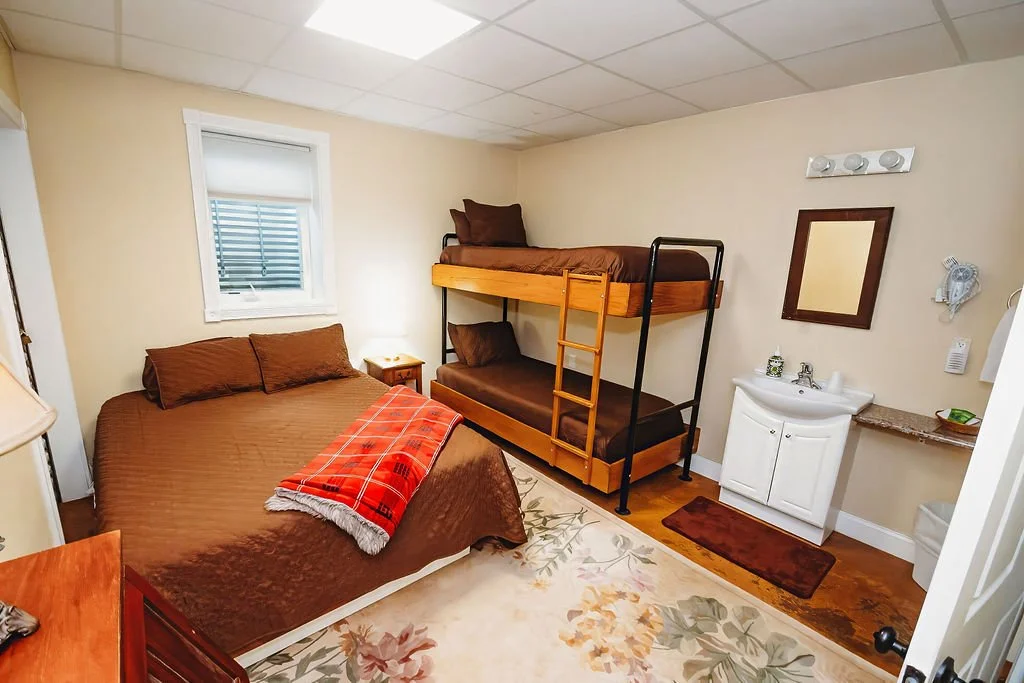 A small bedroom with a full-sized bed covered in brown bedding, a red blanket at the foot, a nightstand with a lamp, a set of bunk beds with brown bedding, a small white sink, a mirror, and a window with blinds.