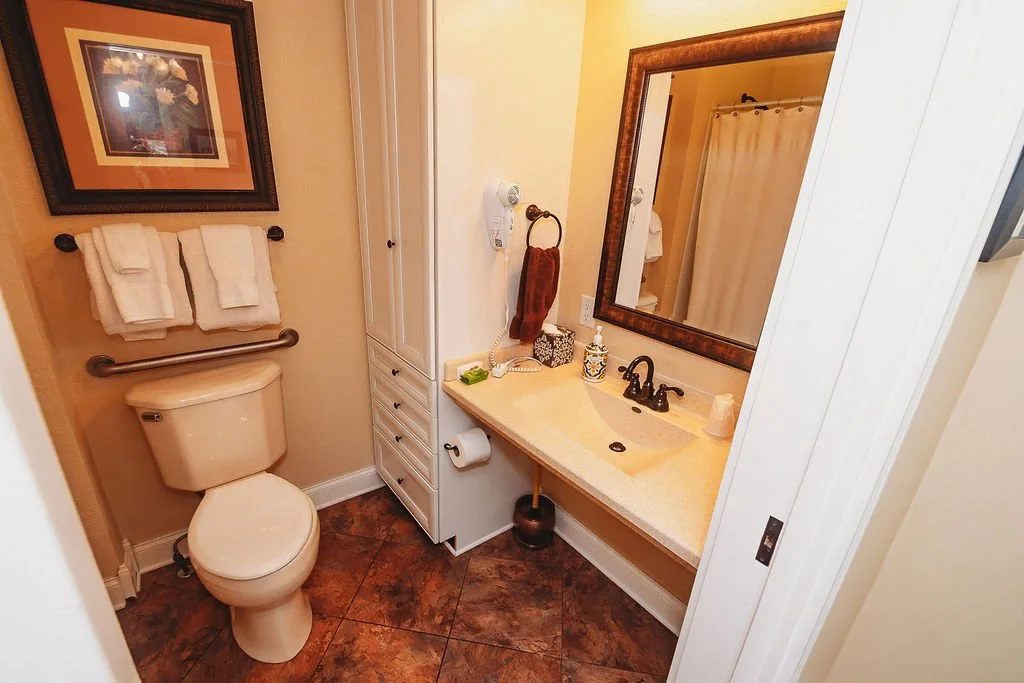 Small bathroom with a toilet, a sink with a large mirror, and a medicine cabinet. White towels are hanging over the toilet tank, and there is a framed picture on the wall above. The bathroom has a brown tiled floor and beige walls.