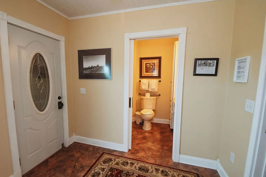 Interior view of a house showing a doorway leading to a bathroom with a toilet. The room has beige walls, white trim, a framed picture on the wall, and a decorative rug on a tiled floor.