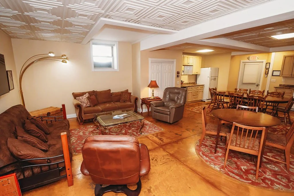 Cozy living room with brown sofas, a leather armchair, a glass coffee table, a patterned area rug, dining table with chairs, and a small kitchen area in the background.