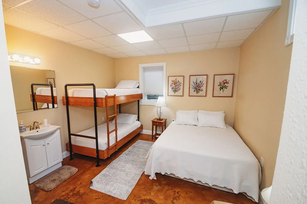 A small bedroom with a bunk bed on the left, a twin bed on the right, a small nightstand with a lamp, a window with blinds, and three framed floral prints on the wall.