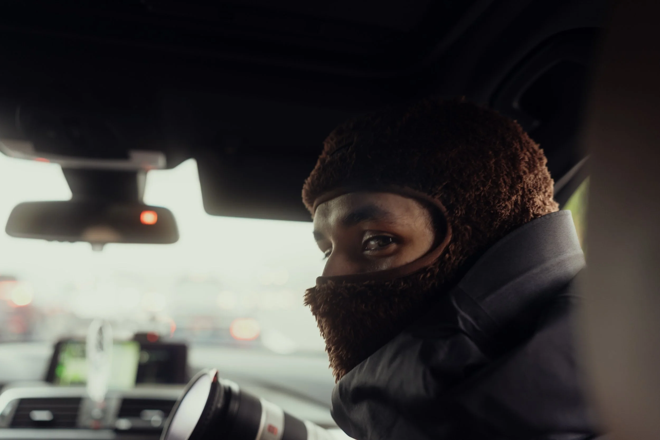 A person looking back over their shoulder while sitting in the driver's seat of a car, wearing a brown furry hat and a matching brown face mask.