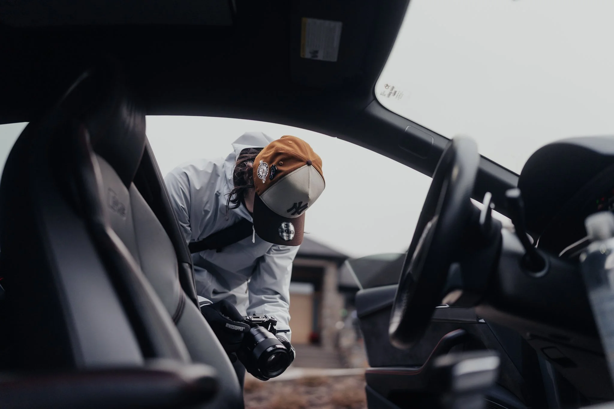 Person in a white jacket and a brown and beige baseball cap leaning into the open door of a vehicle, holding a camera.