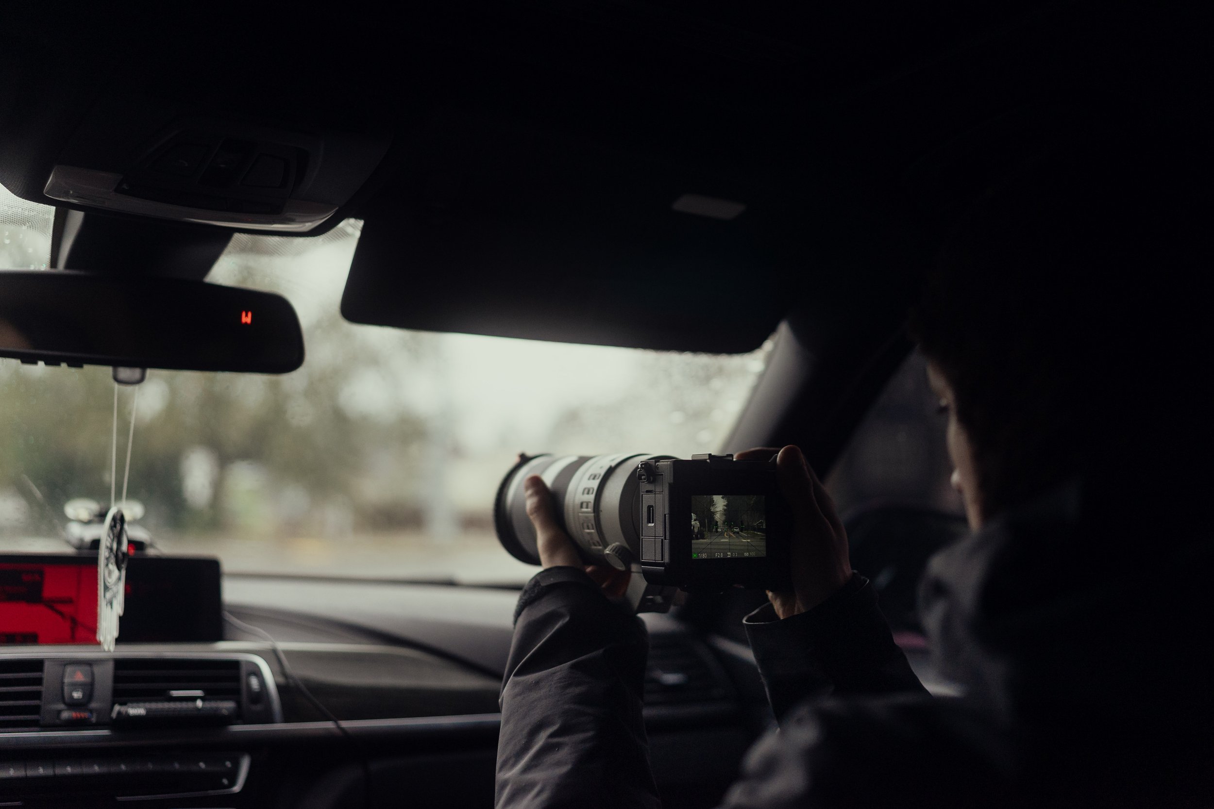 Person inside a car holding a camera with a telephoto lens, photographing or filming through the windshield on a rainy day.