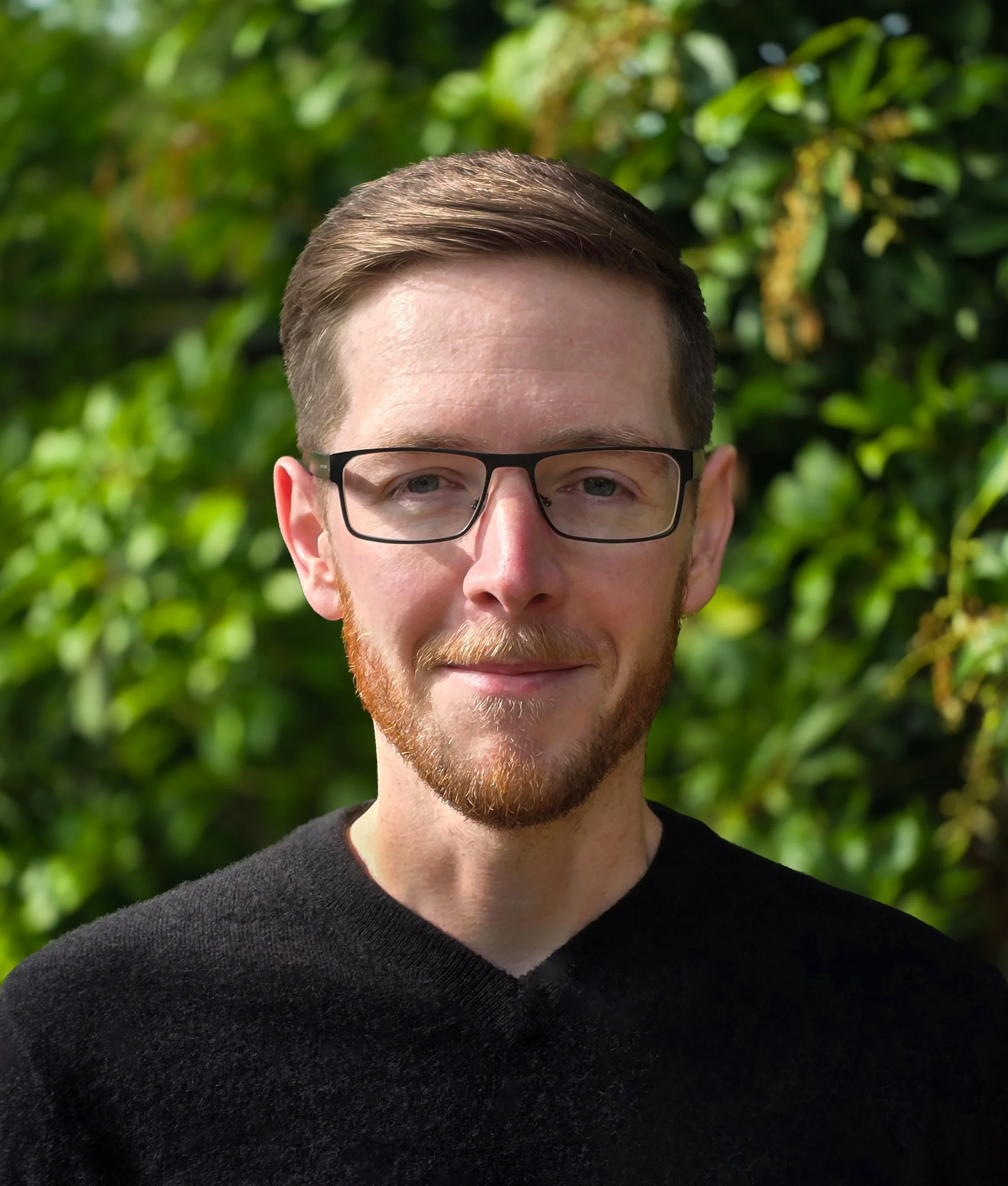 A man with glasses and a beard standing outdoors in front of green leafy bushes.