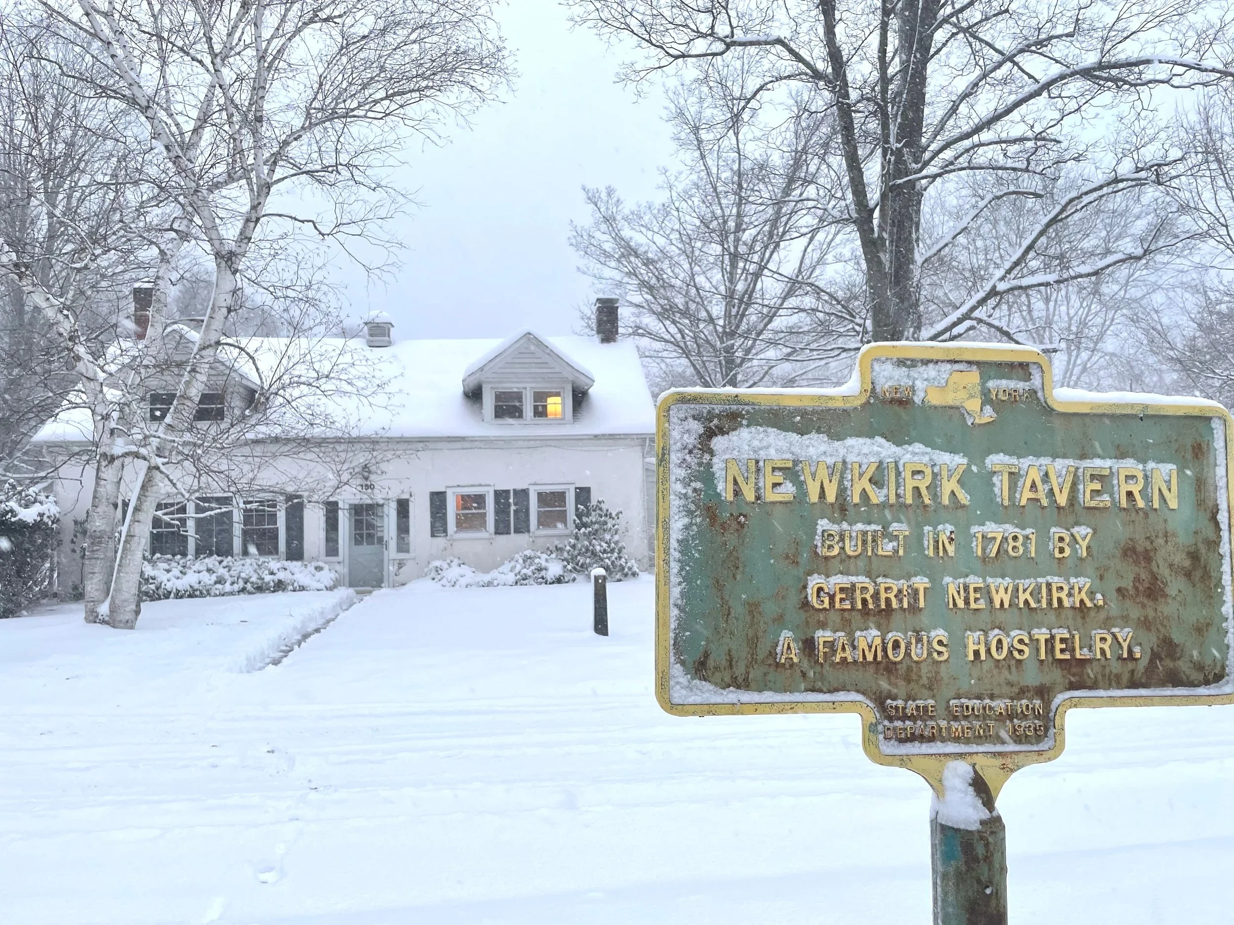 Snowy scene with a historic sign for Newkirk Tavern, built in 1781 by Gerrit Newkirk, in front of a snow-covered house with leafless trees.